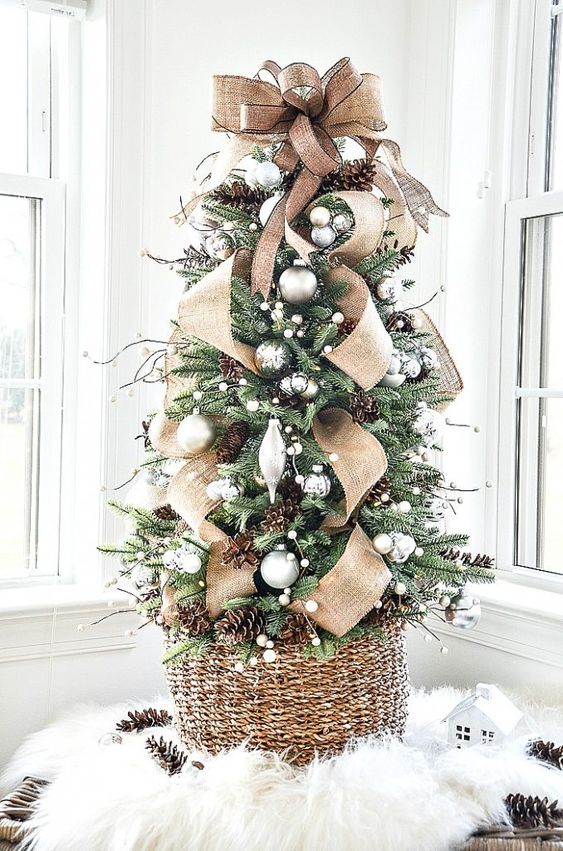 a gorgeous tabletop Christmas tree with silver and metallic ornaments, pinecones, twigs and a large burlap bow on top