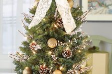 an elegant tabletop Christmas tree with gold glitter ornaments, snowy pinecones, lights, willow and a large white and gold bow on top