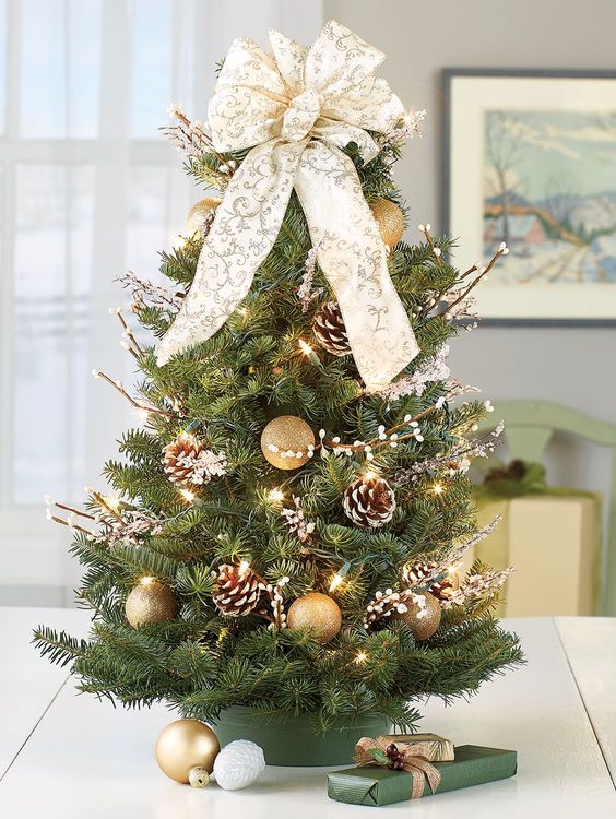 an elegant tabletop Christmas tree with gold glitter ornaments, snowy pinecones, lights, willow and a large white and gold bow on top