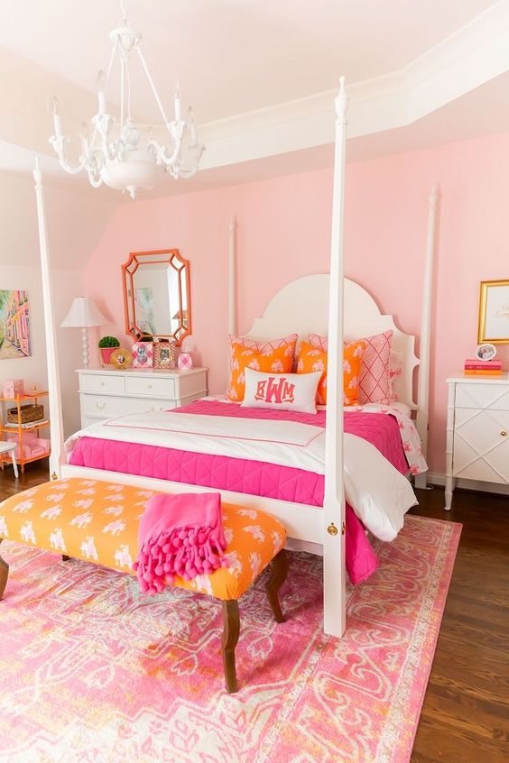 a bright pink girl's room with a vintage bed with colorful bedding, a yellow bench with a pink blanket, a white chandelier and a pink printed rug