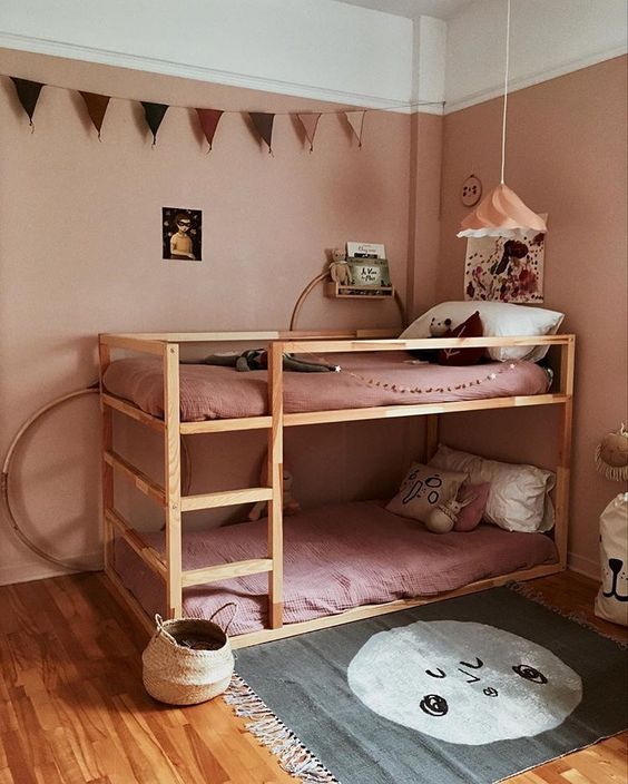 a dusty pink kid's room with dusty pink walls and bedding, an IKEA Kura bunk bed, a printed rug, a garland and baskets