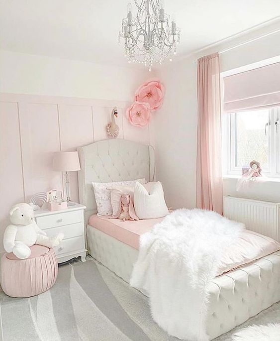 a light pink and white girl's bedroom with a white upholstered bed with pink and bedding, a white nightstand, a pink pouf and a crystal chandelier