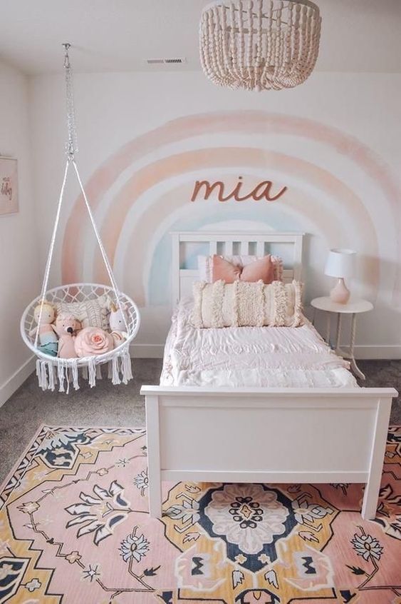 a lovely girl's room with a rainbow accent wall, a white bed and a nightstand,  pendant chair, a pink printed rug is a cute and cozy space
