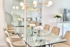 a modern and catchy dining table with a glass tabletop and a black curved base, with beige chairs and a gold chandelier over the table