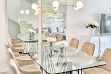 a modern and catchy dining table with a glass tabletop and a black curved base, with beige chairs and a gold chandelier over the table