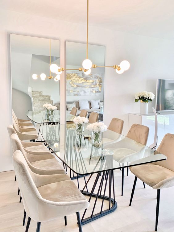 a modern and catchy dining table with a glass tabletop and a black curved base, with beige chairs and a gold chandelier over the table
