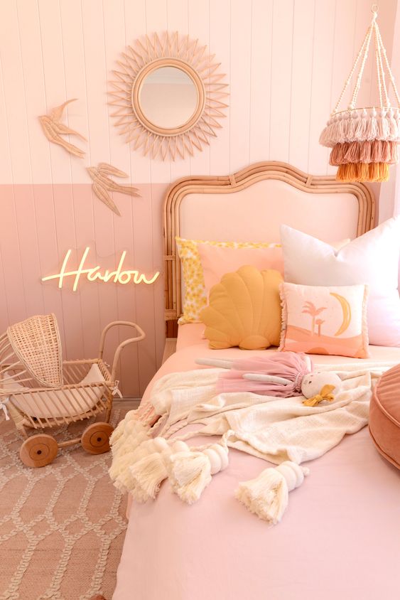 a pink and blush girl's room with color block walls, a blush bed, blush bedding, mellow yellow pillows, a tassel chandelier and a wicker baby carriage