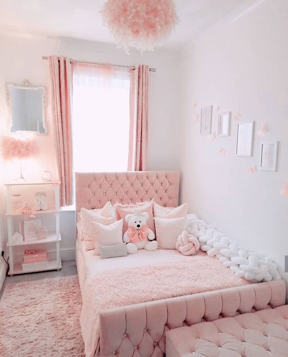 a pink and white girl's room with a pink upholstered bed and a chest bench, a white shelving unit, fluffy pink lamps