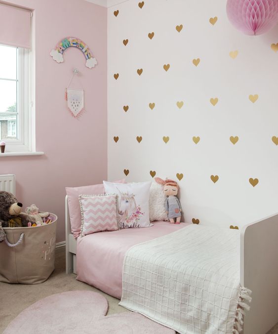 a pretty girl's bedroom with a light pink wall, a gold heart accent wall, a white bed with pink and white bedding, a basket with toys