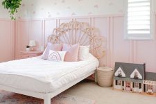 a pretty kid’s room with light pink panels, pink printed wallpaper, a wicker bed with pink and white bedding, a dollhouse