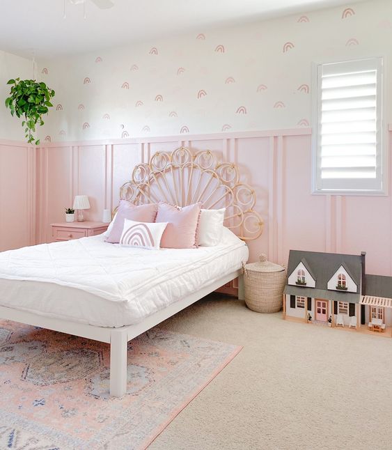 a pretty kid's room with light pink panels, pink printed wallpaper, a wicker bed with pink and white bedding, a dollhouse
