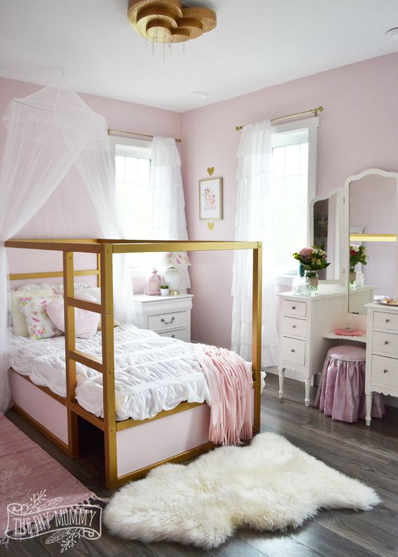 a shabby chic pink girl's bedroom with pink walls, gold canopy beds with pink and white bedding, a vintage vanity and a pink stool