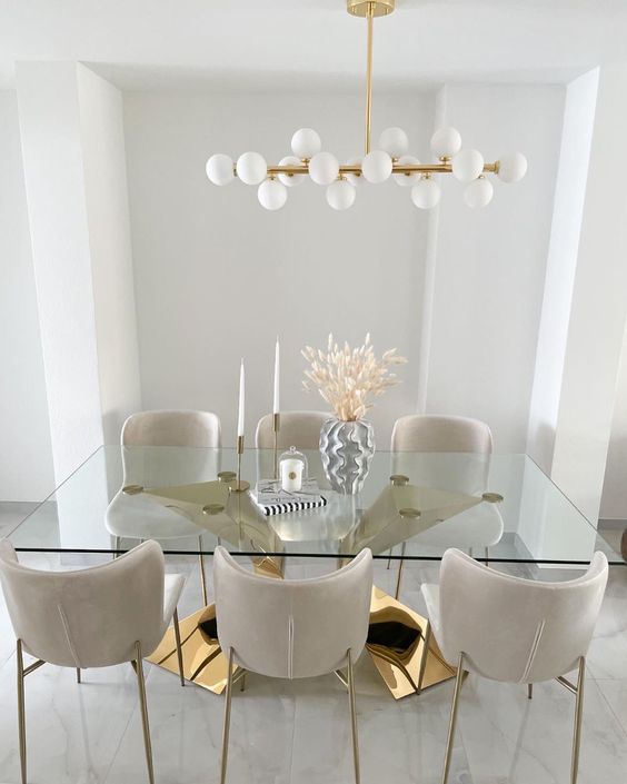 a sophisticated dining room with a large polished gold base and a glass tabletop, creamy chairs and a mid century modern chandelier