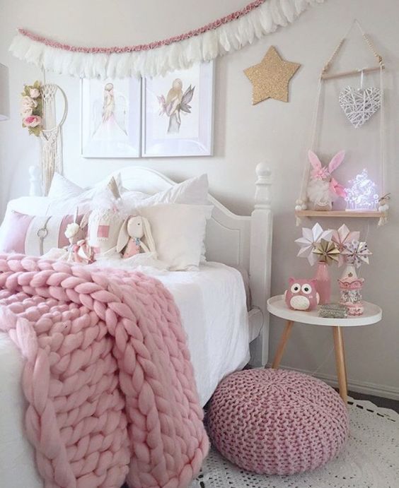 a white bedroom with vintage and modern white furniture, pink blankets and a pouf, a tassel garland and pink toys