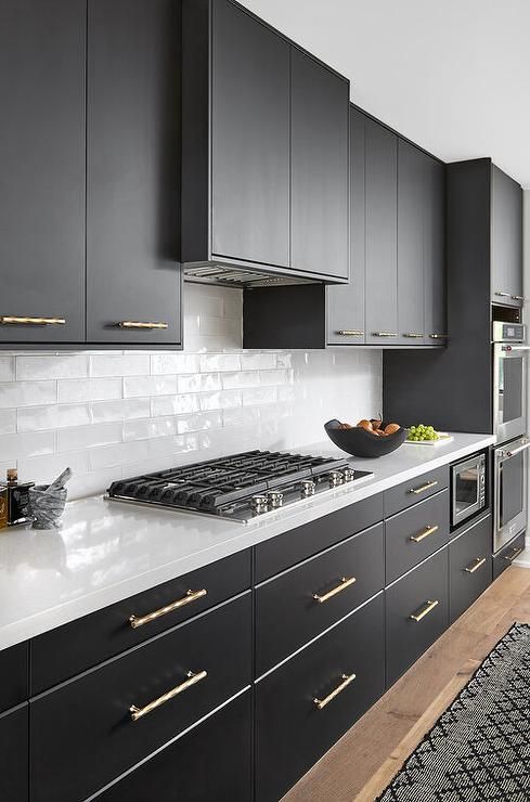 a chic black and white kitchen with sleek and matte black cabinets, white countertops, white skinny tiles on the backsplash and gold fixtures