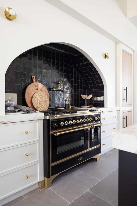 a chic vintage kitchen with white shaker cabinets, black Zellige tiles, a vintage black cooker and gold fixtures and touches