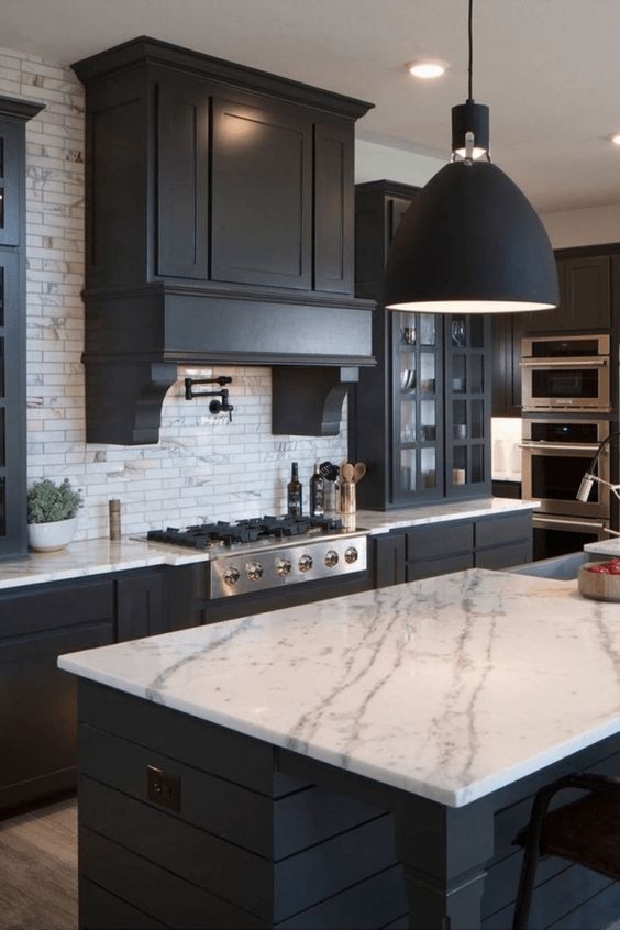 a modern black and white farmhouse kitchen with sleek matte cabinets, a large vintage hood, white marble countertops and a white marble tile backsplash, black lamps