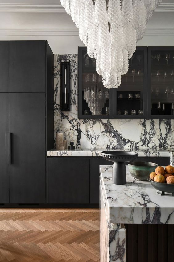 a sophisticated black and white kitchen with matte black cabinets and white marble countertops and a backsplash plus a stunnign crystal chandelier