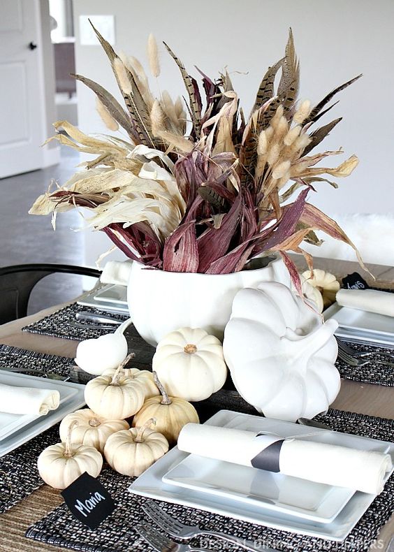 a Thanksgiving centerpiece of a white soup bowl with wheat, feathers, corn husks and some white pumpkins around it