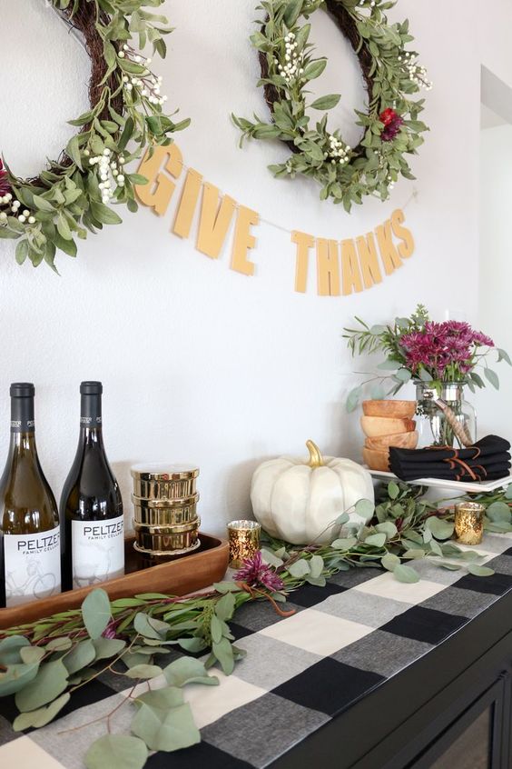 a Thanksgiving food and drink station with a plaid runner, a white pumpkin, pink florals and wreaths over it