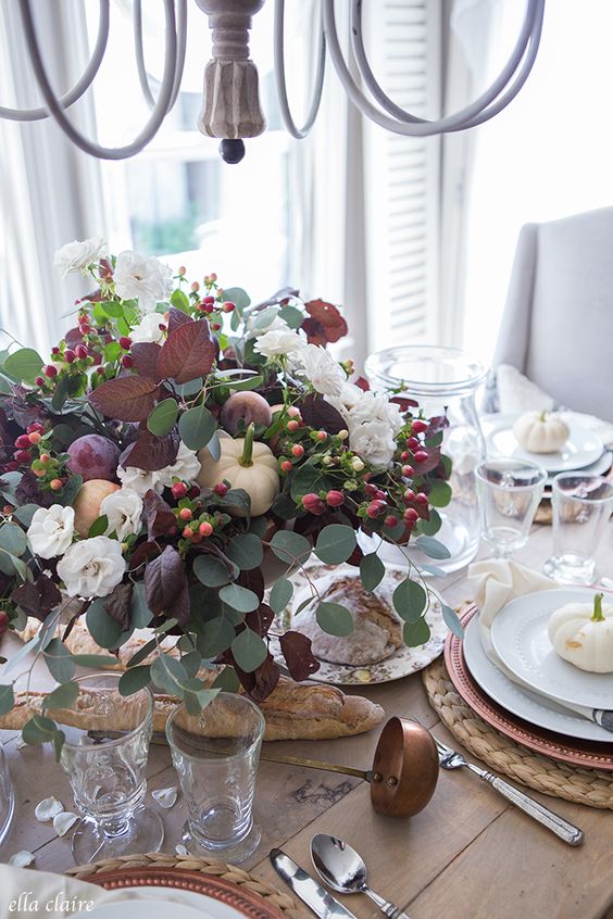 a beautiful Thanksgiving centerpiece of greenery, dark foliage, berries, white blooms and some fresh fruits