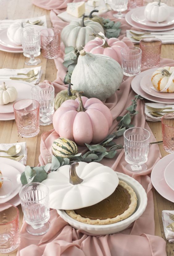 a beautiful pastel Thanksgiving tablescape with a blush runner, plates and glasses, some natural pumpkins and greenery