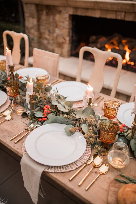 a chic Thanksgiving table with a greenery and berry runner, candles, amber glasses and woven chargers plus gold cutlery
