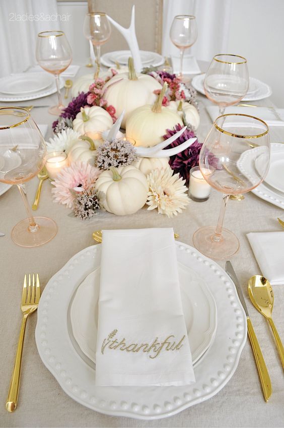 a glam Thanksgiving table setting with a centerpiece of white pumpkins and neutral and purple blooms, candles, gold cutlery and blush glasses