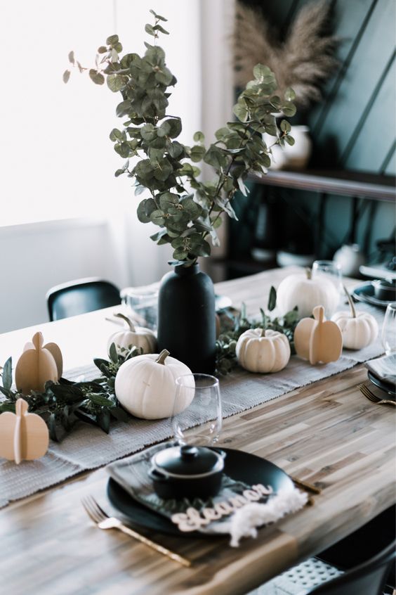 a modern Thanksgiving table with white pumpkins - real and cardboard ones, a greenery arrangement in a black vase and black plates