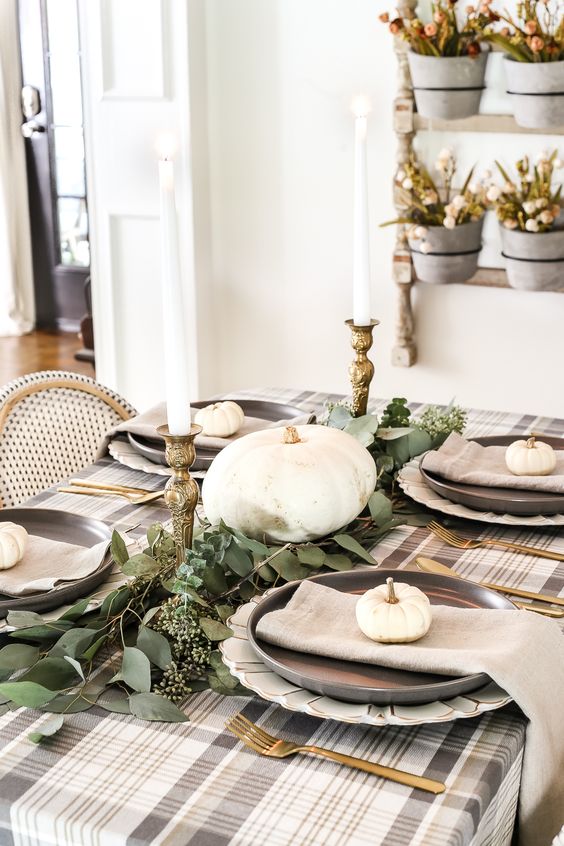 a neutral low-key Thanksgiving tablescape witha plaid tablecloth, grey napkins, a greenery runner, white pumpkins and candles