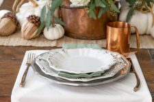 a neutral rustic Thanksgiving table with a plaid runner, a white napkin, a copper mug, a bucket with hydrangeas and roses and acorns