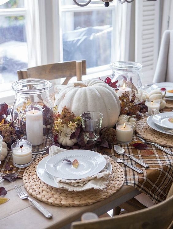 a neutral rustic Thanksgiving tablescape with a plaid runner, woven chargers, pumpkins, dark foliage and candles around