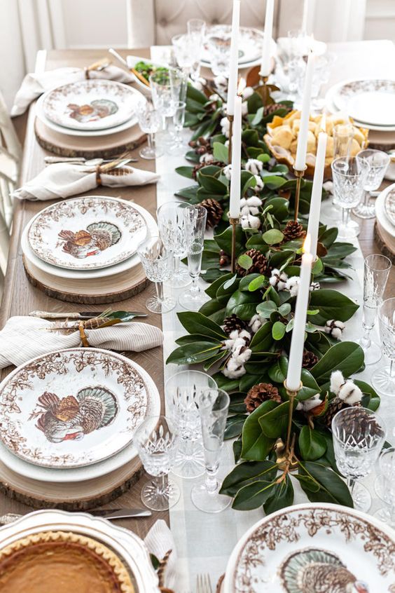 a traditional Thanksgiving tablescape with a magnolia leaf runner, cotton, pinecones, candles and printed porcelain plus wood slice chargers