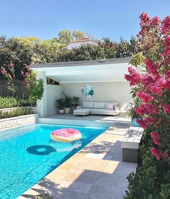 a contemporary space with a blue pool, neutral stone tiles, a neutral sofa with pillows, potted plants and blooms