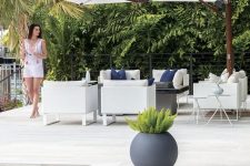 a lovely modern outdoor space with a pool, a white stone deck, white seating furniture and a parasol plus greenery around