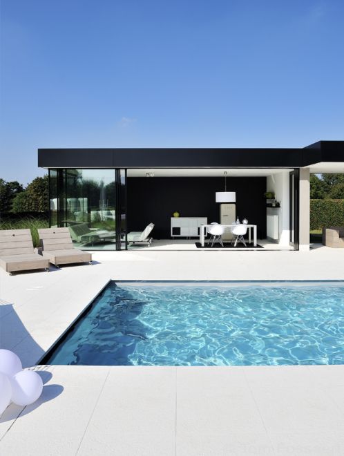 a minimalist outdoor space with a blue pool, a white stone tile deck, woodne loungers and a small yet cool pool house