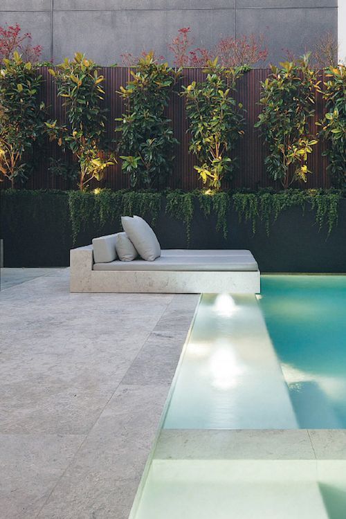 a minimalist outdoor space with a large lit up pool, a neutral stone deck and a built-in bed with neutral bedding