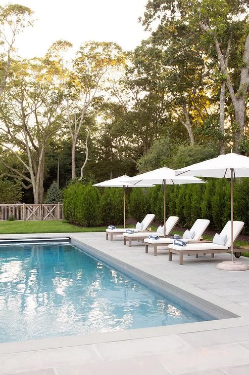 a neutral elegant outdoor space with a white stone deck, a pool, several white loungers under umbrellas and greenery around