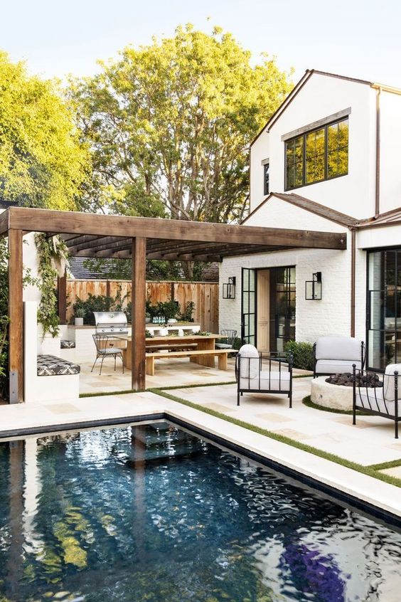 a neutral outdoor space with a pool, a white stone tile deck, greenery, chairs around a fire pit and a dining space under a roof