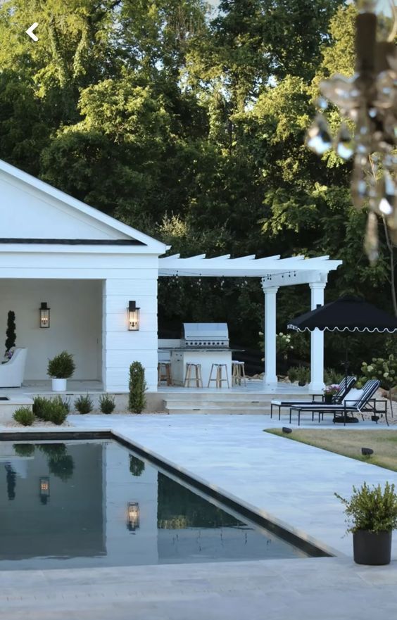 a peaceful modern outdoor space with a pool, a stone tile deck, black and white loungers and a parasol, an outdoor kitchen