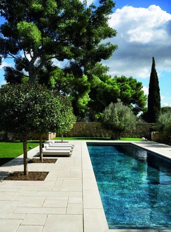 a refined outdoor space with a pool, a neutral stone deck, some loungers and some planted trees is a very cool space