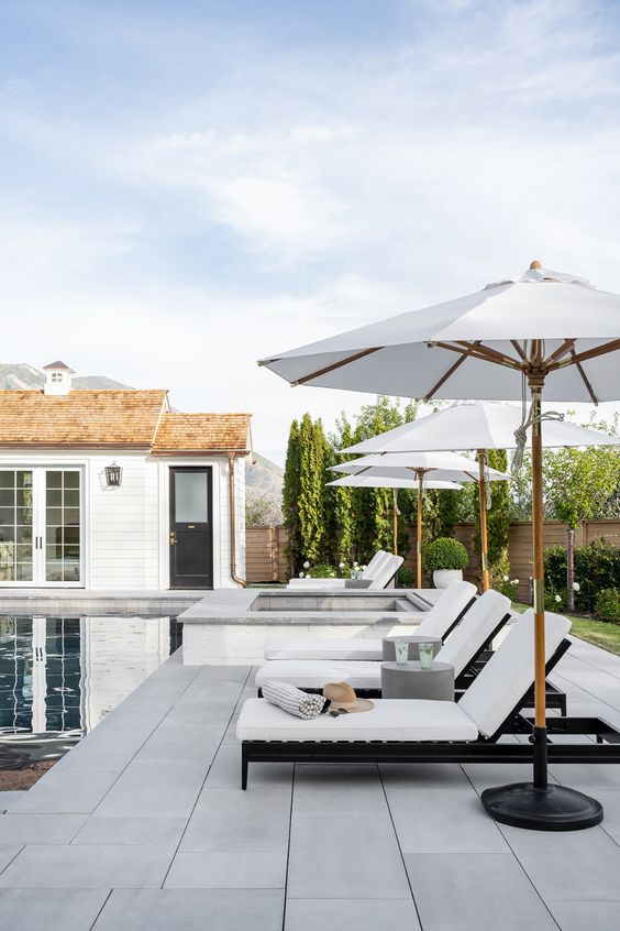 a sleek modern outdoor space with a pool, grey stone tiles, a jacuzzi, white loungers and parasols, some greenery and trees