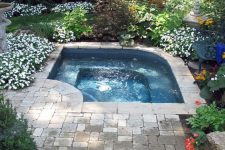 a small and lovely outdoor space with a small pool, a stone deck and lots of blooms around, with greenery and green lawn