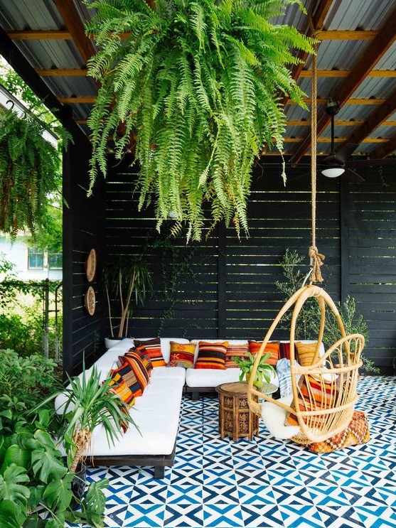 a beautiful tropical patio with a roof and greenery hanging down, with an L-shaped sofa with colorful pillows, a rattan chair and a mosaic tile floor