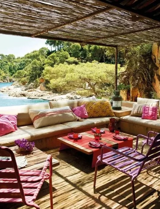 a bright and colorful outdoor living room with neutral upholstered benches, colorful pillows, bright stools and textiles plus a sea view