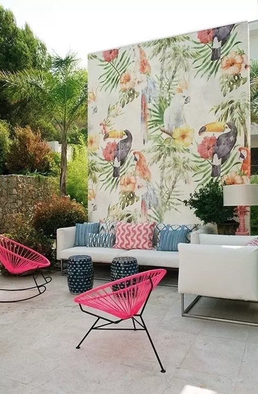 a colorful tropical outdoor living room with a white sofa and colorful pillows, hot pink chairs, a bright watercolor wall and little stools