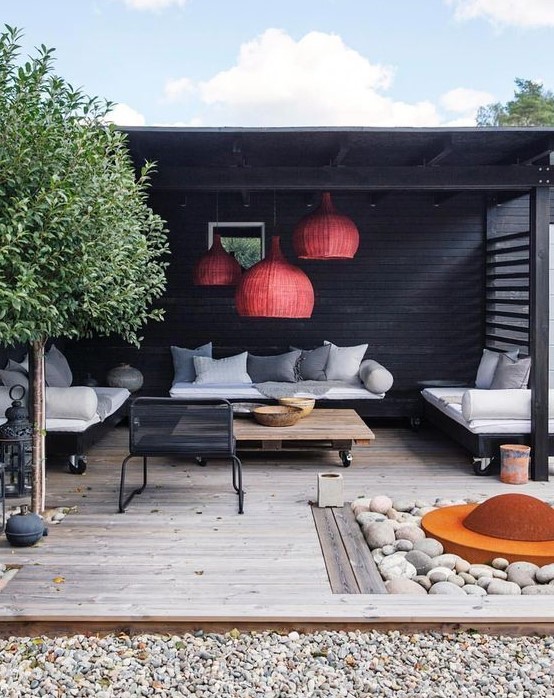 a modern outdoor living room with red wicker lamps, modern furniture on casters, a fire pit with pebbles around and a tree