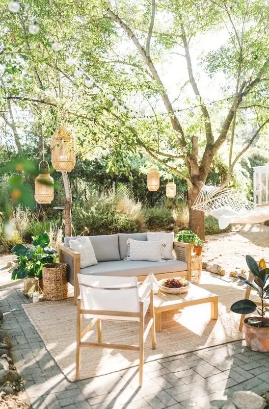 an outdoor living room with a grey sofa, neutral chairs, a low coffee table, lanterns hanging over the space, potted plants
