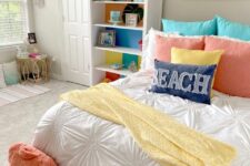a bright beach teen girl room with a white storage unit, a bed with colorful bedding, coral pillows on the floor, a rattan table and a beach artwork