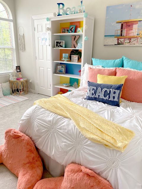 a bright beach teen girl room with a white storage unit, a bed with colorful bedding, coral pillows on the floor, a rattan table and a beach artwork